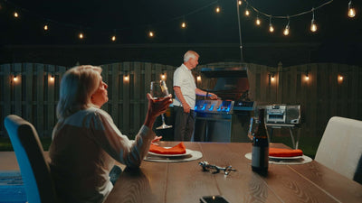 Two people sitting at a table outdoors with string lights and a grill in the background.