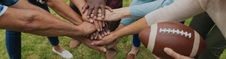 Group of people with diverse skin tones stacking their hands together, holding a football on grass.