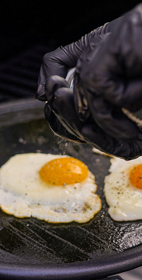 Person wearing black gloves cooking eggs on a grill