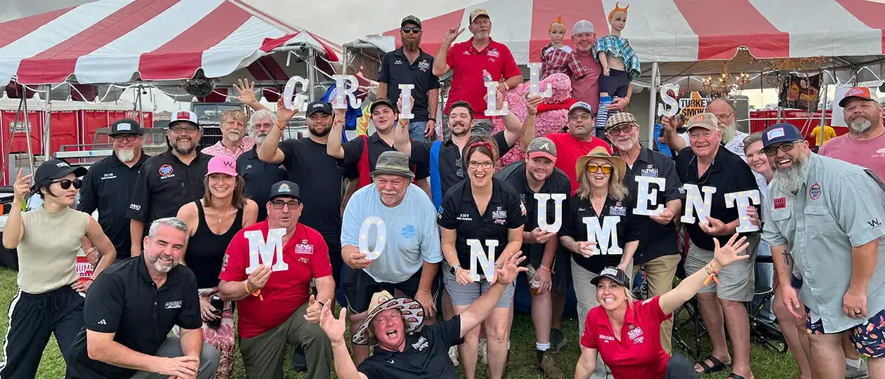 Group of people holding letters to spell 'MONUMENT GRILLS' at an outdoor event with a red and white striped tent.