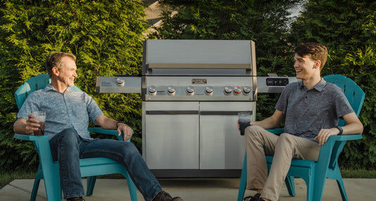 Two men sitting on teal chairs in front of a large barbecue grill outdoors.