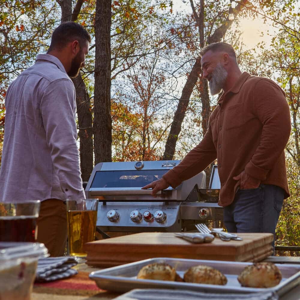 Two men cooking outdoors with Mesa 415BZNG | Stainless Natural Gas Grill in a scenic forest setting.