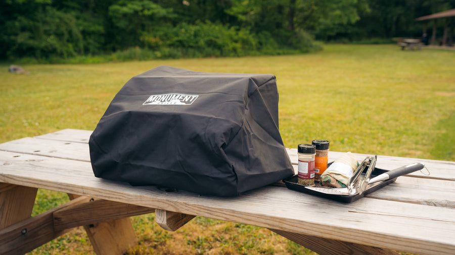 Covered tabletop grill on a picnic table beside seasonings and grilling utensils.