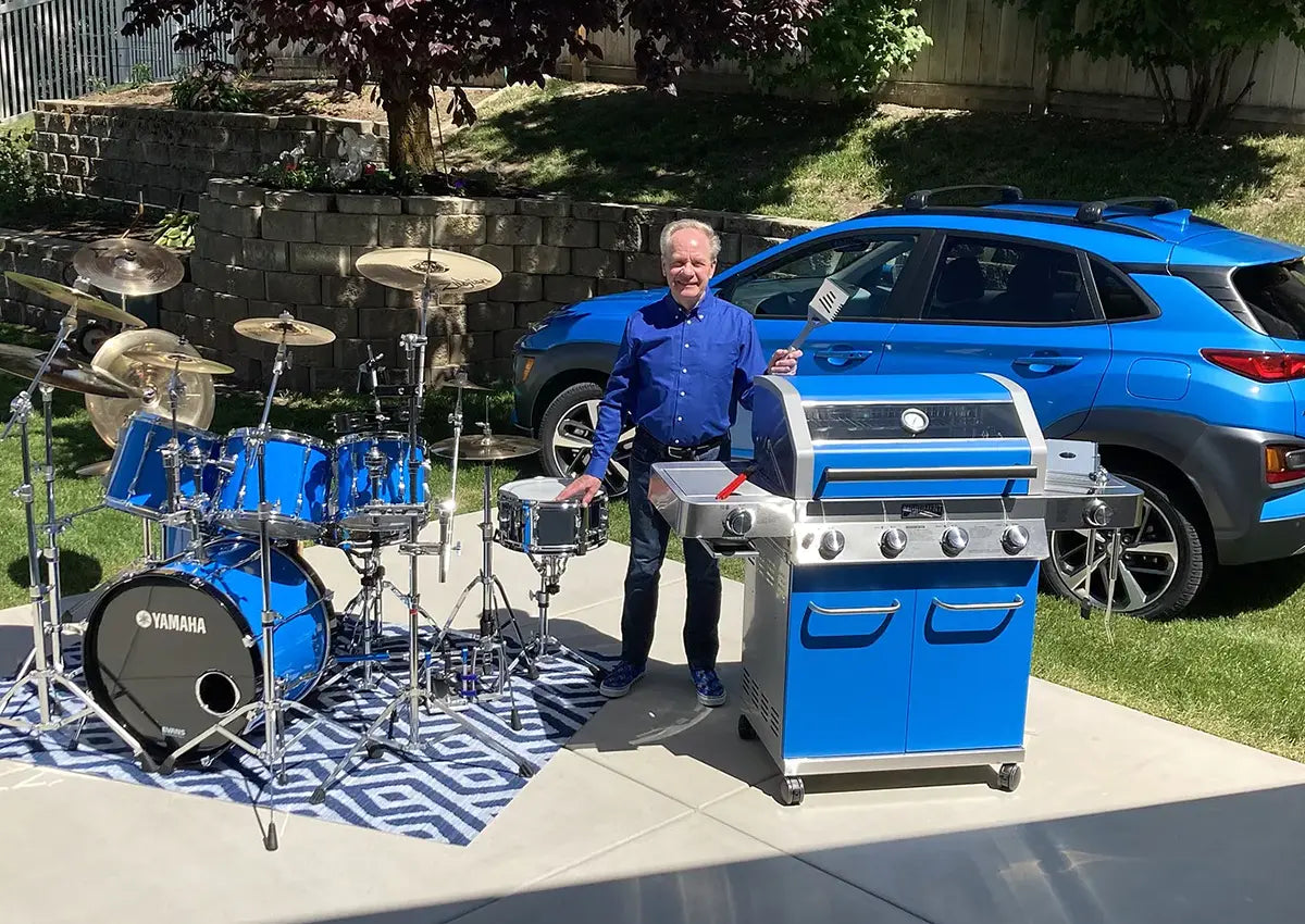 Man standing next to a blue Yamaha drum set and a matching barbecue grill in a driveway.