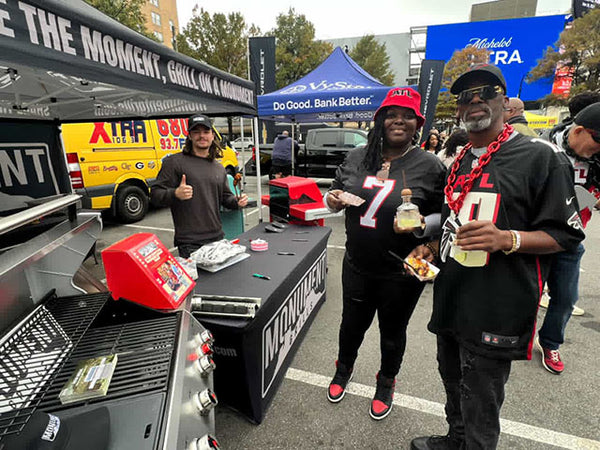 Two men standing next to a grill with promotional items and branding in the background.