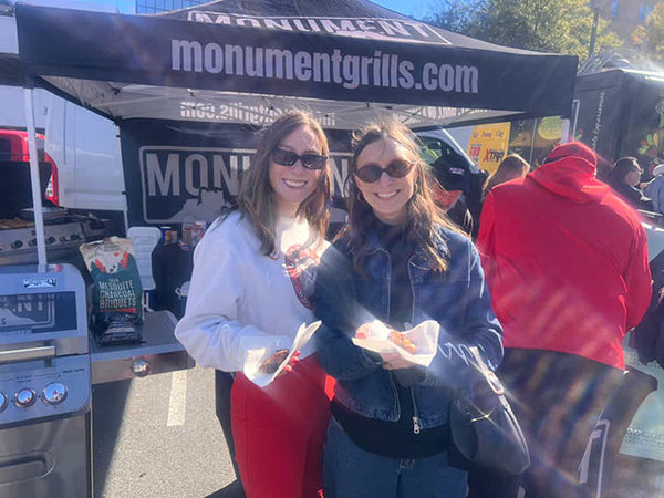 Two women holding food under a Monument Grills canopy at an outdoor event.