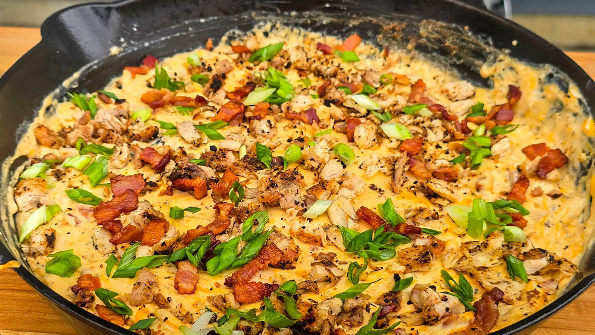 A close-up shot of a creamy cheesy casserole or skillet dish topped with bacon bits, chopped green onions, and mushrooms, served in a black cast-iron skillet on a wooden surface.