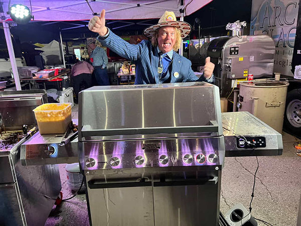 Person in a cowboy hat standing next to a large barbecue grill at an outdoor event.