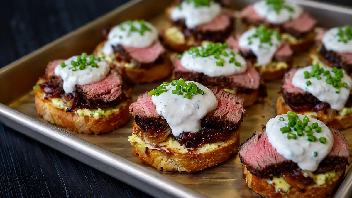 Close-up of several gourmet appetizers on a metal baking sheet, featuring sliced pink steak, caramelized onions, a creamy white sauce, and chopped chives on toasted bread.