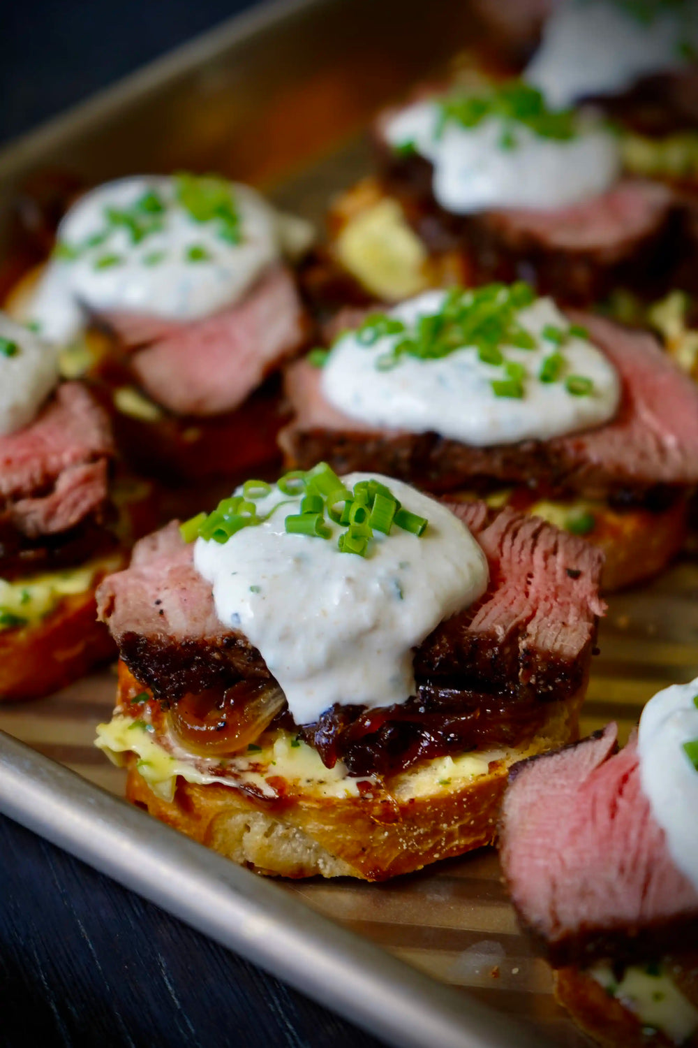 Close-up of several gourmet appetizers on a metal baking sheet, featuring sliced pink steak, caramelized onions, a creamy white sauce, and chopped chives on toasted bread.