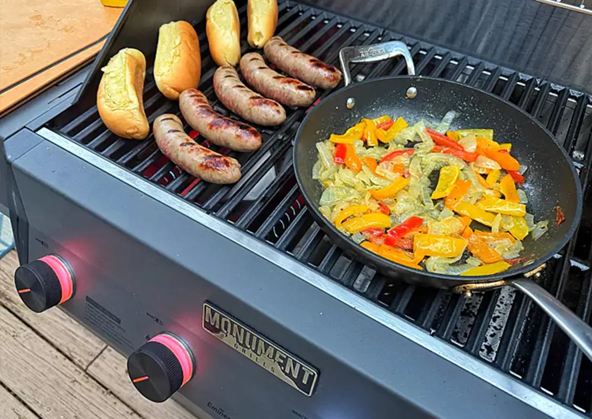 Grill with sausages and buns on one side and a pan of vegetables on the other, on a wooden surface.