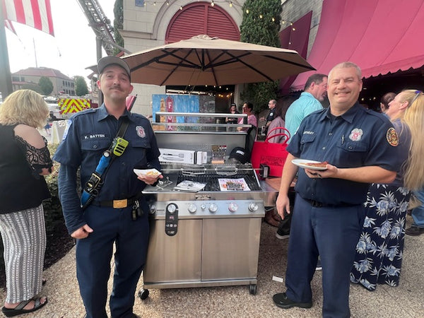 Two firefighters standing next to a portable grill with food, surrounded by people and a pink building.