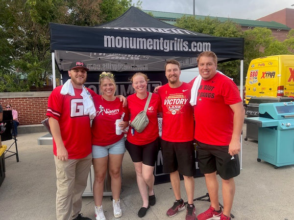 Group of people in red shirts standing in front of a 'monumentgrills.com' tent.