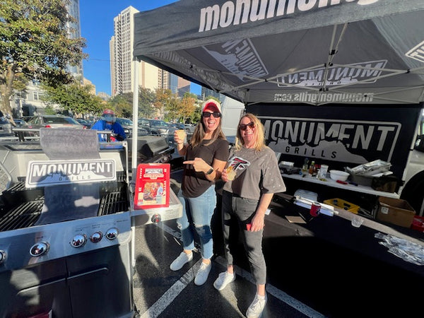 Two women standing next to a Monument Grills grill and tent in an outdoor setting.