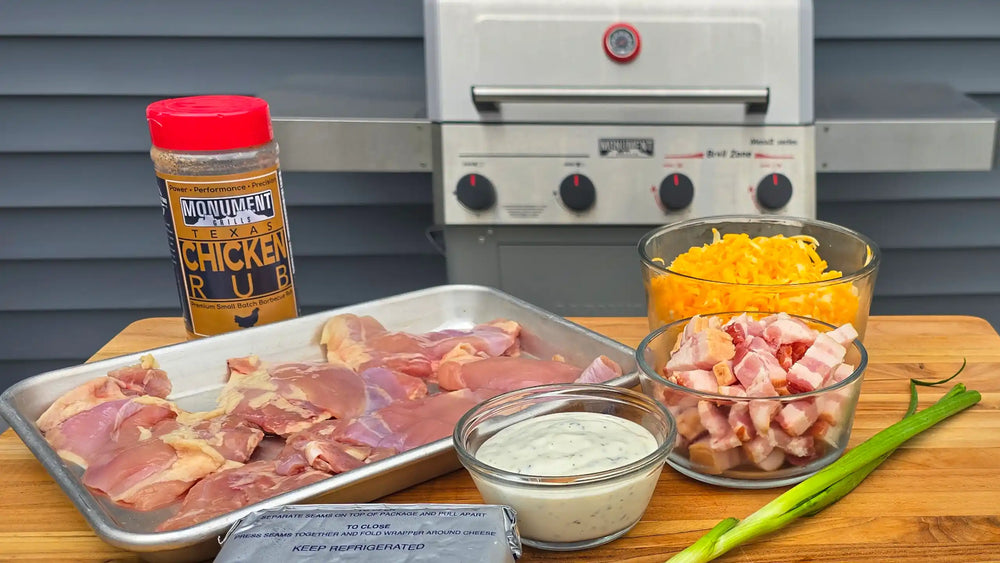Ingredients arranged on a wooden cutting board in front of a grill: raw chicken thighs on a tray, a bottle of Monument Chicken Rub seasoning, shredded cheddar cheese, chopped bacon, a small bowl of white sauce, green onions, and a stick of butter.
