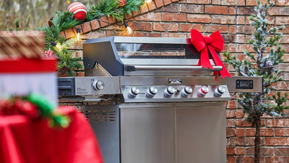 Stainless steel grill with a red bow outdoors against a brick wall with Christmas decorations.