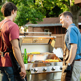 Two men in aprons smiling and chatting while grilling corn and steak on a backyard grill.