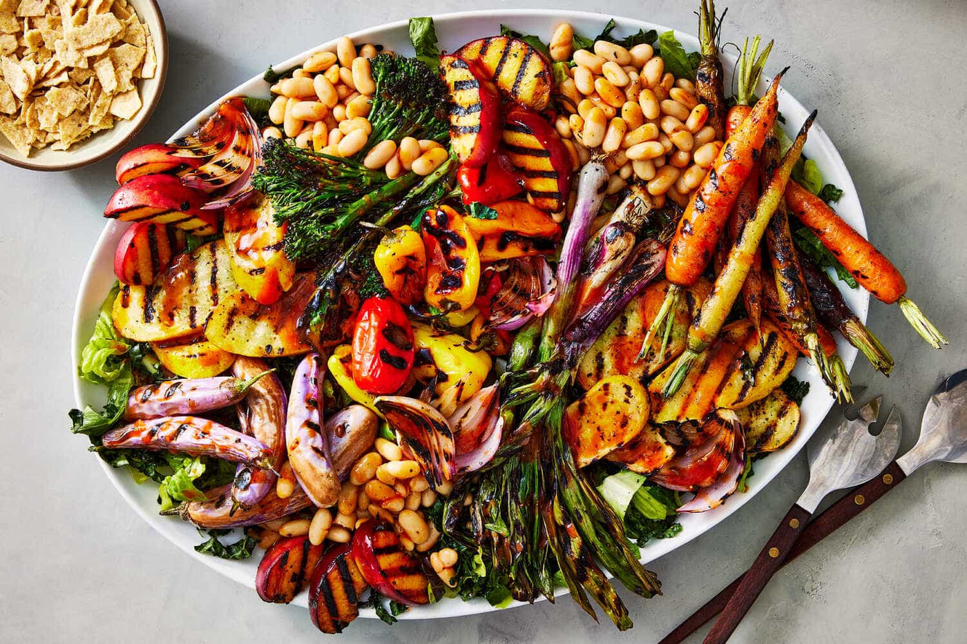 Platter of grilled vegetables and beans on a gray surface with a side of tortilla chips.