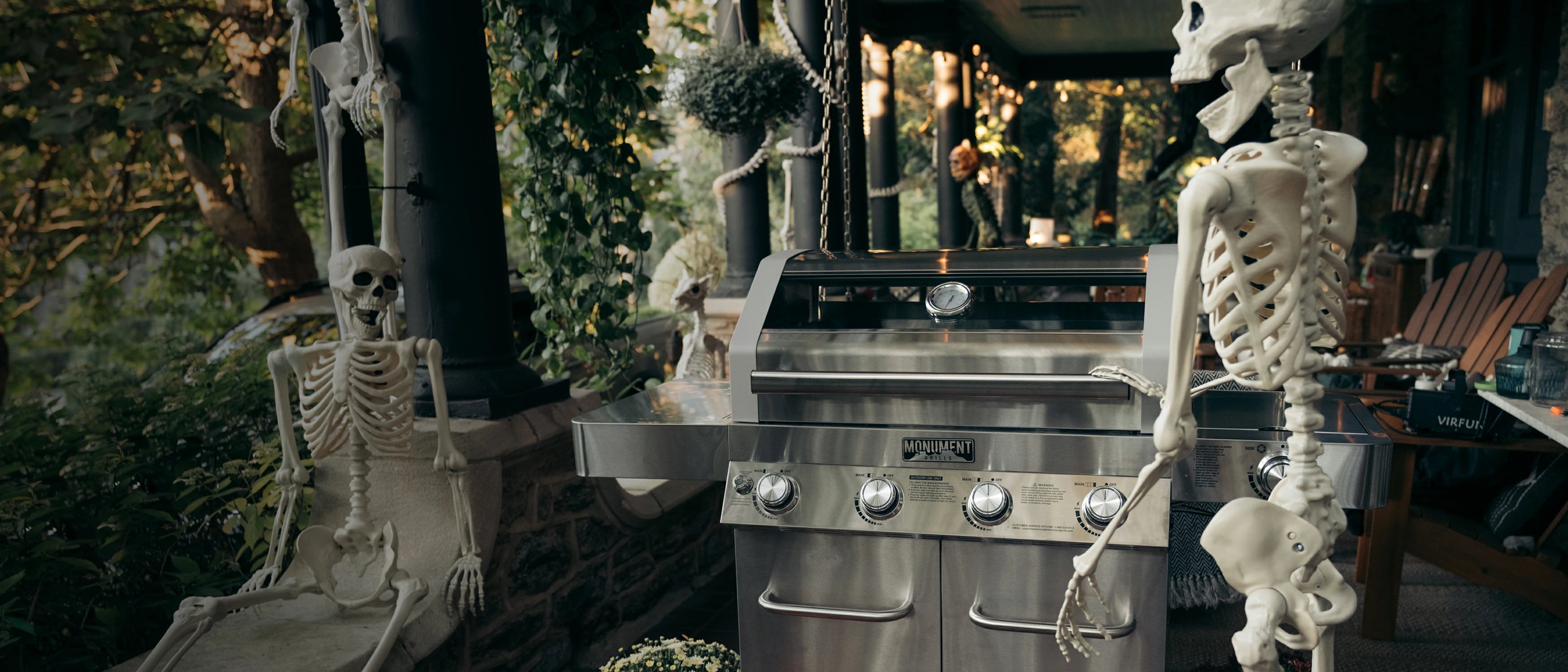 Two skeletons sitting on either side of a barbecue grill in an outdoor setting with greenery.