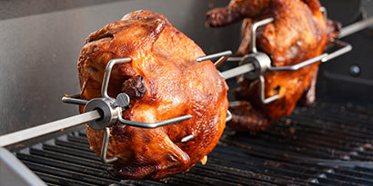 Two roasted chickens on rotisserie skewers over a grill.